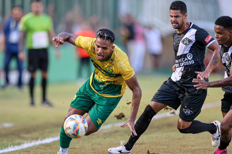 Fotografia de uma partida de futebol em campo de grama natural. Em primeiro plano, um jogador do Cuiabá, vestindo camisa amarela e calções verdes, está em movimento dinâmico tentando controlar a bola com o corpo inclinado. Ele é marcado de perto por dois jogadores adversários que vestem uniformes pretos com detalhes brancos. A ação levanta um pouco de terra do gramado, e o fundo mostra outros jogadores e torcedores de forma desfocada.