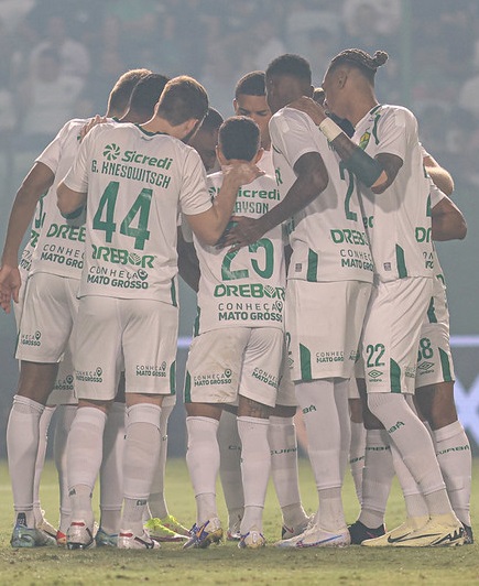 Jogadores do Cuiabá reunidos em um círculo apertado no campo, de costas para a câmera, abraçados pelos ombros em um momento de união antes ou depois de uma partida. É possível ver os nomes e números nas camisas brancas