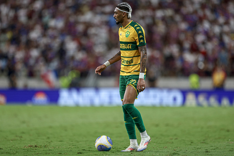 jogador Derik do Cuiabá Esporte Clube durante uma partida. Ele é um homem negro com o cabelo preso em tranças curtas e loiras, vestindo o uniforme titular amarelo com listras verdes horizontais, calções verdes e meiões verdes. Ele está parado no gramado com o pé esquerdo próximo à bola, olhando atentamente para o lado. O fundo mostra as arquibancadas de um estádio lotado e desfocado.