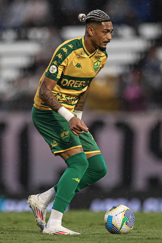 Close-up de ação de Derik do Cuiabá. Ele está em uma postura curvada, conduzindo a bola com o pé direito. É possível ver detalhes das tatuagens em seus braços e o acabamento platinado de suas tranças. Ele usa chuteiras brancas com detalhes coloridos. O ambiente sugere um jogo noturno com iluminação artificial intensa e fundo levemente esfumaçado.