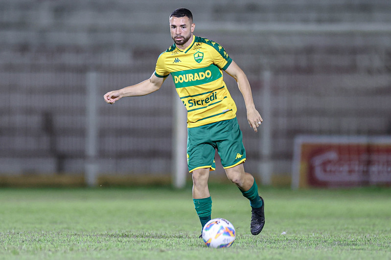 "Fotografia colorida do jogador Léo Ataíde, do Cuiabá, durante uma partida de futebol. Ele é um homem pardo, de barba aparada e cabelo curto, vestindo o uniforme principal do clube: camisa amarela com detalhes verdes e a inscrição 'DOURADO', calções verdes e meiões verdes. Ele está em movimento no gramado, conduzindo a bola com o pé direito. O fundo mostra as arquibancadas de um estádio de forma desfocada."