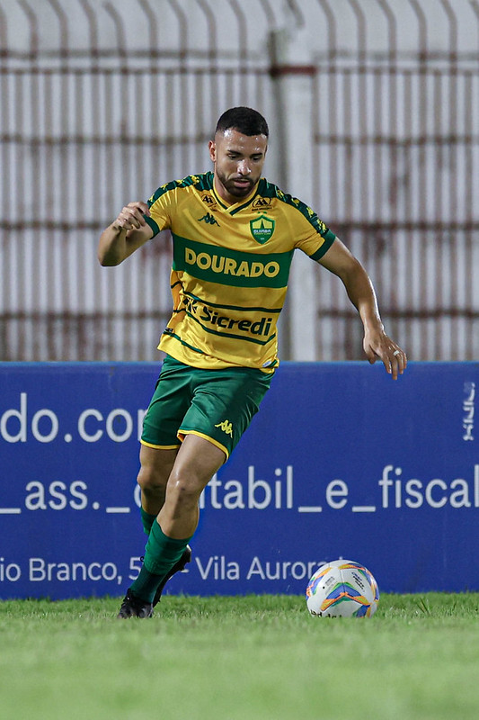 "Léo Ataíde, lateral do Cuiabá, em ação durante jogo noturno. O jogador aparece focado, inclinando o corpo para a esquerda enquanto controla a bola de futebol no gramado. Ele veste o uniforme oficial amarelo e verde da marca Kappa, com patrocínio da Sicredi. Ao fundo, uma placa de publicidade azul e uma grade branca delimitando o campo."