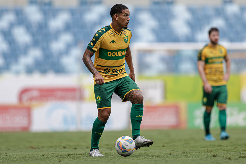 O volante Denilson em campo, visto de corpo inteiro em um ângulo lateral. Ele utiliza chuteiras brancas e controla uma bola de futebol oficial sobre o gramado. O uniforme da marca Kappa destaca o patrocínio master "Dourado" e o selo da "Sicredi". Outro jogador do Cuiabá aparece ao fundo, fora de foco, observando a jogada.
