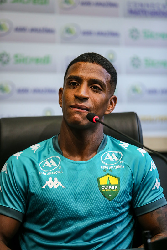O jogador Max, de pele retinta e cabelos curtos, aparece sentado em uma cadeira preta de coletiva, posicionado em frente a um microfone. Ele veste uma camisa de treino verde-azulada com os logos da Kappa, Agro Amazônia e o escudo do Cuiabá.