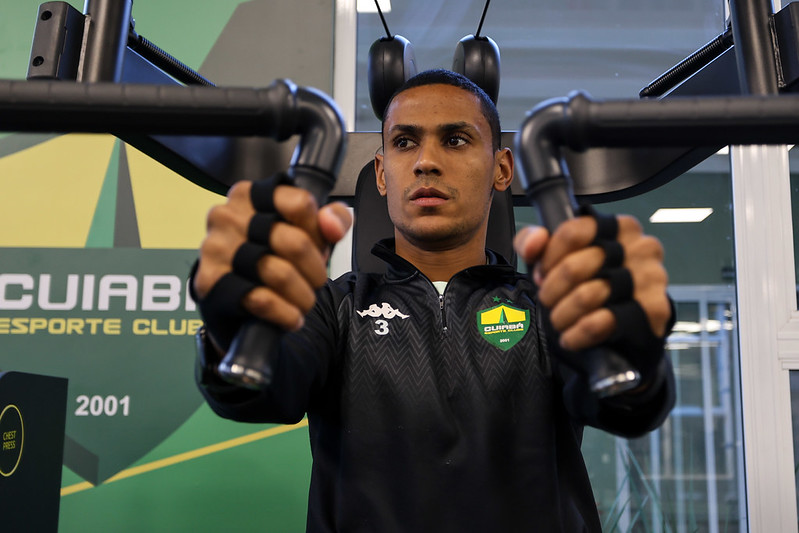 "Fotografia em plano médio de Bruno Alves, jogador do Cuiabá, realizando um exercício de supino em uma máquina de musculação. Ele é um homem pardo, de cabelos bem curtos, com expressão de foco e concentração. Veste um agasalho de treino preto com o escudo do clube e o número 3 no peito. Suas mãos usam faixas protetoras pretas enquanto seguram as alavancas do equipamento. Ao fundo, uma parede verde com o logotipo do Cuiabá Esporte Clube."