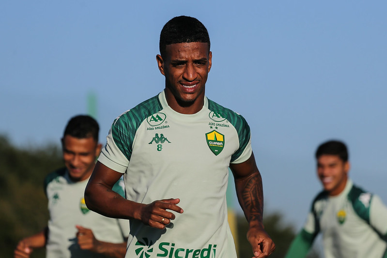 Max aparece em destaque correndo no campo durante o dia. Ele veste o uniforme de treino verde claro com o número 8 pequeno no peito. Ao fundo, outros jogadores aparecem desfocados sob um céu limpo.