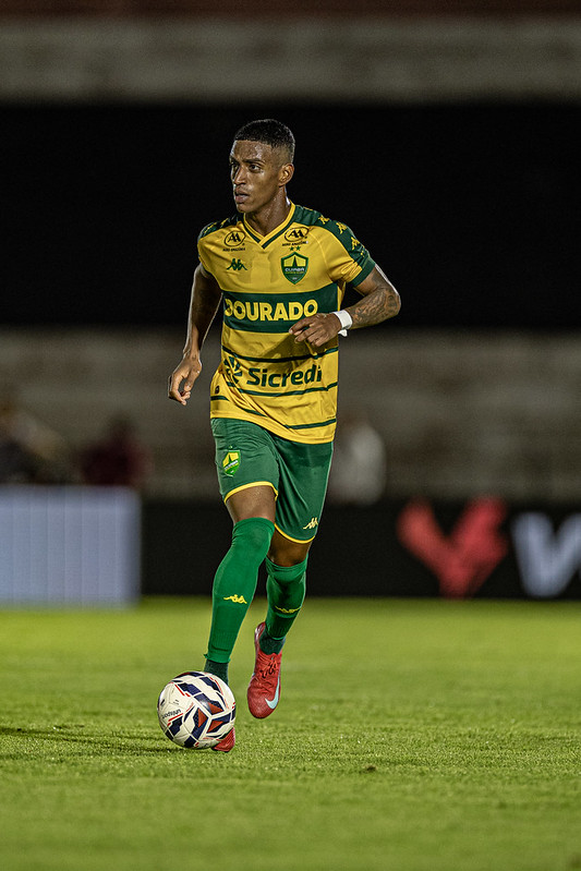 Fotografia em plano médio do jogador Max, do Cuiabá, conduzindo a bola durante uma partida noturna. Ele veste a camisa listrada em verde e amarelo com o patrocínio "Sicredi" em destaque, calções verdes e meiões verdes. Max está focado, olhando para a frente, com o pé direito próximo à bola branca com detalhes em azul e vermelho.
