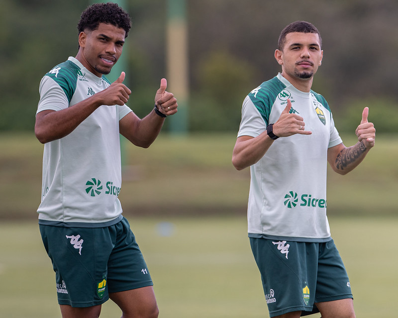 Dois jogadores do Cuiabá Esporte Clube durante treino no CT, vestindo uniforme branco e verde, fazendo sinal positivo com as mãos antes da estreia no Campeonato Mato-grossense.