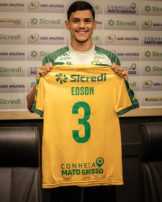 O jogador Edson aparece sorrindo, segurando verticalmente a camisa oficial de jogo do Cuiabá à sua frente.