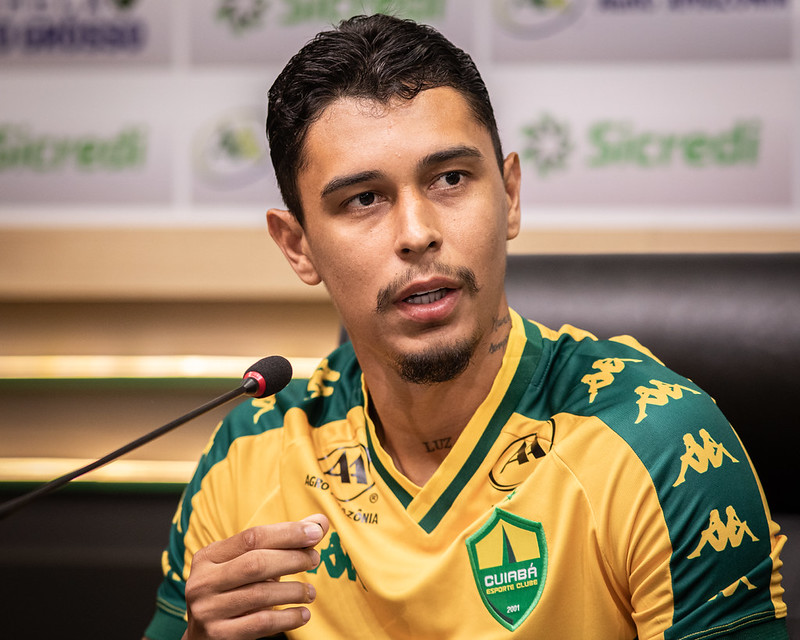 Um close-up de Vitor Mendes sentado durante uma entrevista coletiva. Ele está vestindo o uniforme de treino ou viagem do clube, que é amarelo com o escudo do Cuiabá Esporte Clube no peito e logotipos da marca Kappa nos ombros.