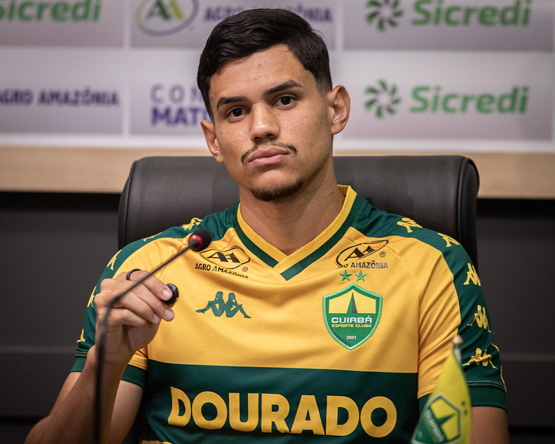 Close-up frontal de Edson sentado em uma cadeira de couro preta, posicionado atrás de um microfone durante uma coletiva de imprensa.