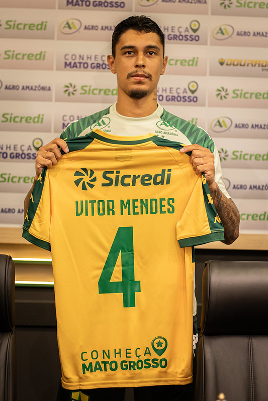 O zagueiro Vitor Mendes aparece de pé, segurando a nova camisa oficial do Cuiabá à sua frente. A camisa é amarela com detalhes em verde nas mangas e gola. Nas costas, destaca-se o nome "VITOR MENDES" e o número 4 em verde.