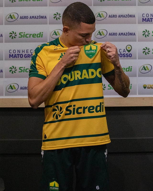 Jogador Marlon vestindo camisa amarela e verde do Cuiabá EC segura e beija o escudo do clube durante apresentação oficial, com painel de patrocinadores ao fundo.