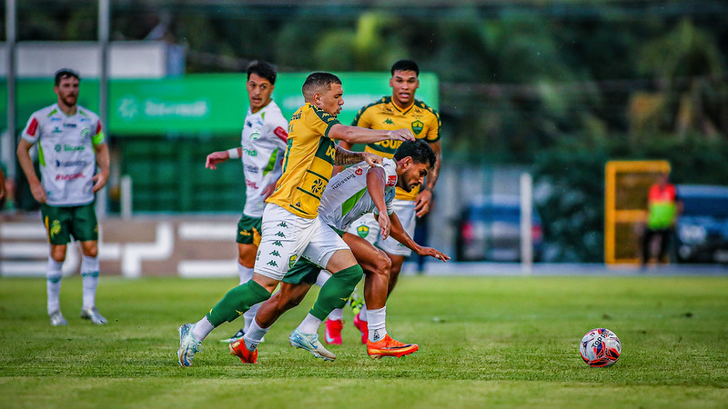 Uma imagem capturada em plano médio mostra uma partida de futebol profissional em um campo gramado durante o dia. No centro da ação, um jogador do Cuiabá (vestindo uniforme amarelo com detalhes verdes e calções brancos) disputa a posse de bola com um jogador do Juventude (vestindo uniforme branco com detalhes verdes).