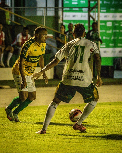 Fotografia em plano médio de um jogo de futebol sob iluminação artificial. Um jogador de costas, vestindo camisa branca com detalhes verdes e o número 11, controla a bola com o pé direito. À sua frente, um jogador do Cuiabá, vestido com a camisa amarela "Dourado", mantém uma postura defensiva. O gramado está bem iluminado e, ao fundo, é possível ver parte da arquibancada e painéis publicitários verdes.