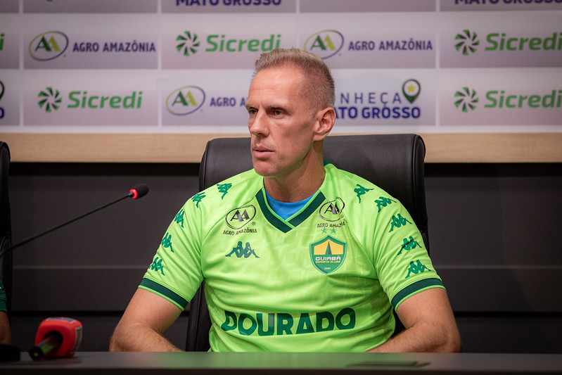 Goleiro Joao Carlos vestindo uma camisa de treino verde neon do Cuiabá Esporte Clube, sentado em uma coletiva de imprensa. Ele olha atentamente para o lado. Ao fundo, um painel com logotipos dos patrocinadores Sicredi e Agro Amazônia.