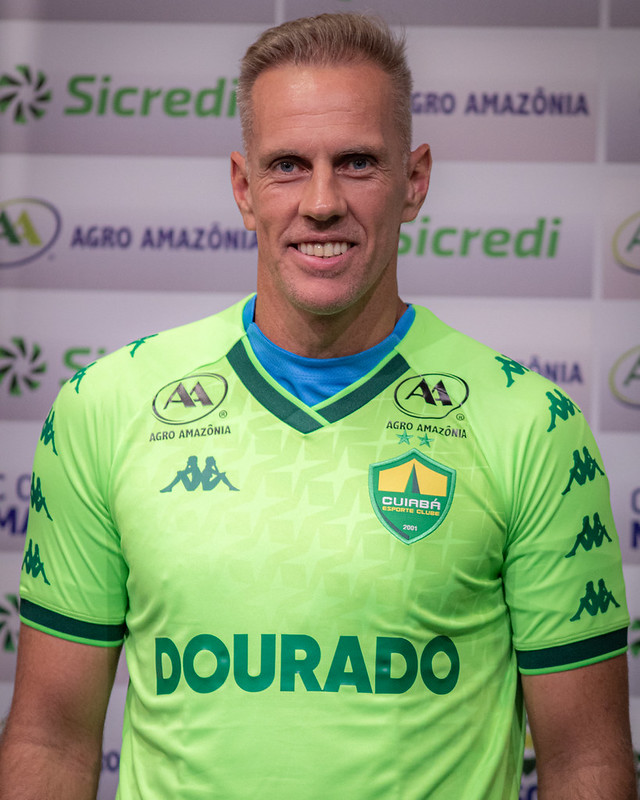 Goleiro Joao Carlos sorrindo para a câmera. Ele veste a camisa verde neon com o escudo do Cuiabá e o patrocínio "Dourado" em destaque. O fundo mostra marcas de patrocinadores de forma repetida.