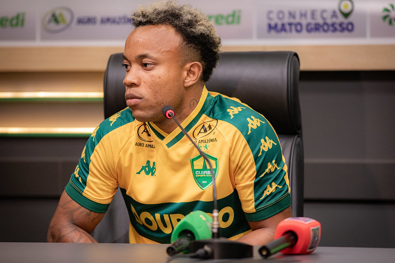 Mateus Santos sentado em uma mesa de coletiva de imprensa. Ele tem pele negra, cabelo curto com o topo descolorido e tatuagens no braço. Veste a camisa oficial de jogo do Cuiabá (amarela com detalhes verdes). À sua frente, microfones de rádio e TV.