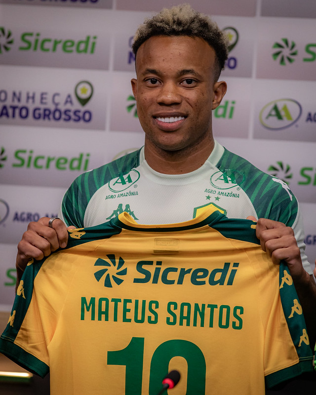 Mateus Santos sorri enquanto segura a camisa número 10 do Cuiabá à sua frente. Na camisa, lê-se "Sicredi", o nome "MATEUS SANTOS" e o número "10" em verde sobre o fundo amarelo. Ele está posicionado em frente ao painel de patrocinadores do clube.