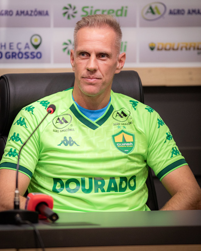 Goleiro Joao Carlos vestindo uma camisa de treino verde neon do Cuiabá Esporte Clube, sentado em uma coletiva de imprensa. Ele olha atentamente para o lado. Ao fundo, um painel com logotipos dos patrocinadores Sicredi e Agro Amazônia.