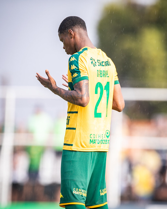 Fotografia de costas do jogador Gabriel, número 21 do Cuiabá, em um campo de futebol. Ele é um homem negro, está com as mãos erguidas em um gesto de oração ou agradecimento e veste o uniforme amarelo e verde. O fundo está desfocado, mostrando a vegetação e a claridade do dia.
