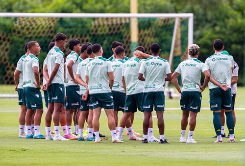 Fotografia em plano médio do grupo de jogadores do Cuiabá reunidos em um círculo no gramado durante um treinamento. Eles estão de costas para a câmera, vestindo uniformes de treino compostos por camisetas verde-claro com o patrocínio da Sicredi e calções verde-escuros. Ao fundo, vê-se uma trave de futebol com rede e uma área de vegetação densa sob luz natural.