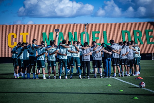 Mostra o grupo de jogadores e a comissão técnica reunidos em um círculo (o tradicional "huddle" ou roda de conversa) no gramado. Ao fundo, destaca-se o muro do centro de treinamento com a inscrição "CT MANOEL DRE..." (referindo-se ao CT Manoel Dresch). O céu está aberto com algumas nuvens, indicando condições ideais para o treino.