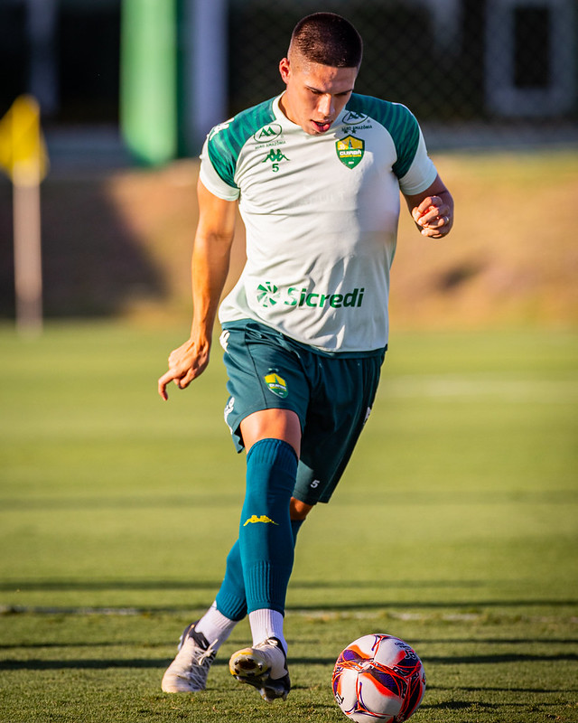 Calebe aparece focado, conduzindo a bola com o pé direito em um gramado bem iluminado pela luz do sol (provavelmente um treino vespertino no CT do clube).