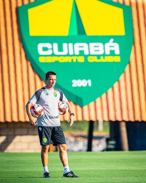 Fotografia do tecnico Eduardo Basrros, em pé no centro do gramado. Ele segura duas bolas de futebol, uma sob cada braço, e veste uma camiseta cinza de treino e calções pretos com o escudo do clube. Ao fundo, destaca-se um grande painel com o escudo oficial do Cuiabá Esporte Clube em verde e amarelo, posicionado sobre uma estrutura de telhado alaranjado.