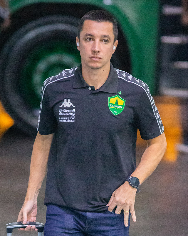 Técnico do Cuiabá caminha pelo corredor da Arena Pantanal usando camisa oficial do clube, com expressão concentrada e fones de ouvido.
