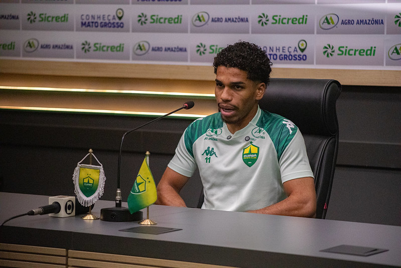 Eliel está sentado à frente de um painel de patrocinadores (backdrops), onde é possível ver marcas como Sicredi, Agro Amazônia e o selo "Conheça Mato Grosso". À sua frente, há microfones e um copo personalizado do clube.