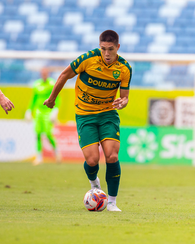 Calebe conduz a bola durante partida do Cuiabá na Arena Pantanal, usando uniforme auriverde, em jogo do Campeonato Mato-grossense sob forte calor.