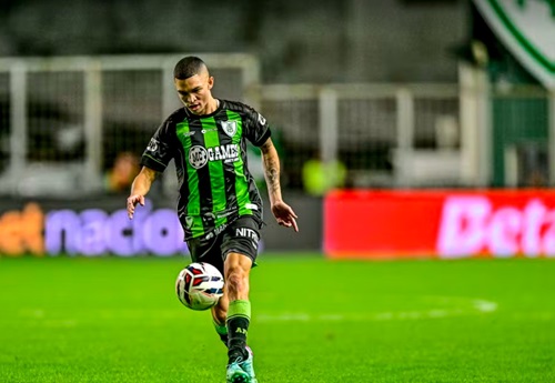 Fotografia de ação do jogador Marlon concentrado durante uma partida noturna. Ele está em movimento, preparando-se para dominar ou chutar a bola de futebol que está no ar à sua frente. Veste o uniforme listrado em verde e preto. O fundo mostra as luzes do estádio e placas de publicidade desfocadas.
