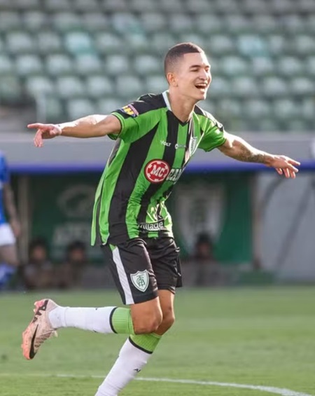 O lateral Marlon aparece sorrindo e correndo pelo gramado com os braços abertos em comemoração. Ele veste o uniforme do América-MG, com listras verticais verdes e pretas, e calções pretos. Ao fundo, as arquibancadas do estádio estão visíveis sob a luz do dia.