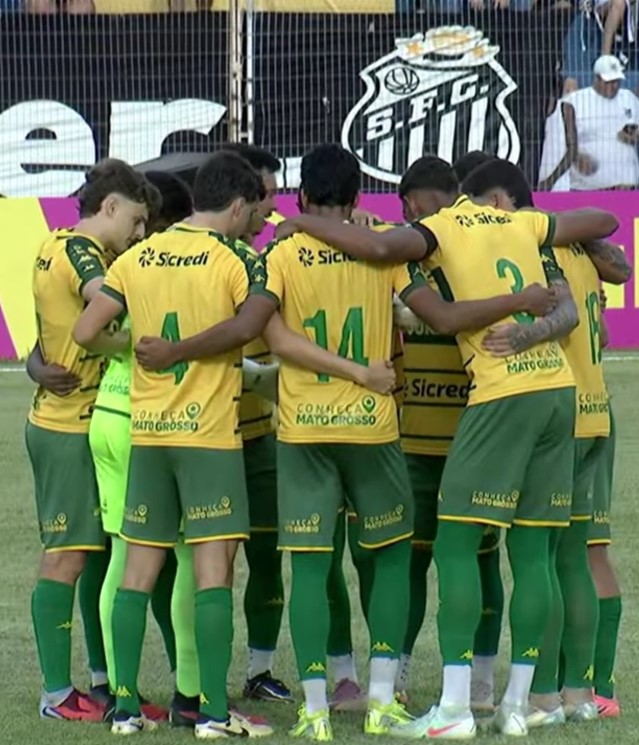 Jogadores do Cuiabá reunidos em roda no gramado durante partida da Copinha 2026, exibindo uniforme amarelo e verde com patrocínio Sicredi, em estádio com escudo do Santos ao fundo.