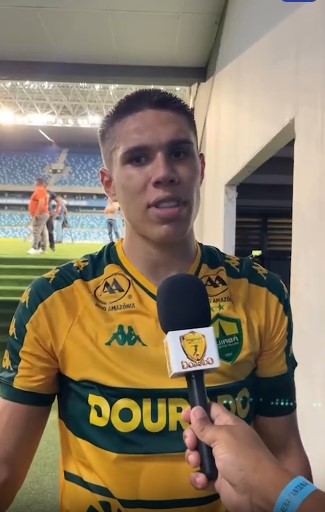 Jogador Calebe do Cuiabá Esporte Clube concede entrevista no gramado da Arena Pantanal após a partida. Atleta veste camisa amarela e verde do Dourado enquanto fala ao microfone com logotipo do clube, com arquibancadas ao fundo.