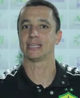 Fotografia de Eduardo Barros, integrante da comissão técnica do Cuiabá Esporte Clube, durante uma entrevista. Ele veste uma camisa polo preta do time e está à frente de um painel com logotipos de patrocinadores como Sicredi e Agro Amazônia.