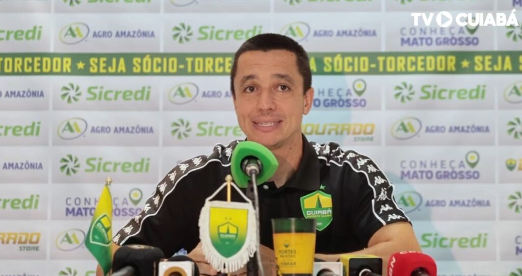 Fotografia de Eduardo Barros, integrante da comissão técnica do Cuiabá Esporte Clube, durante uma entrevista. Ele veste uma camisa polo preta do time e está à frente de um painel com logotipos de patrocinadores como Sicredi e Agro Amazônia.