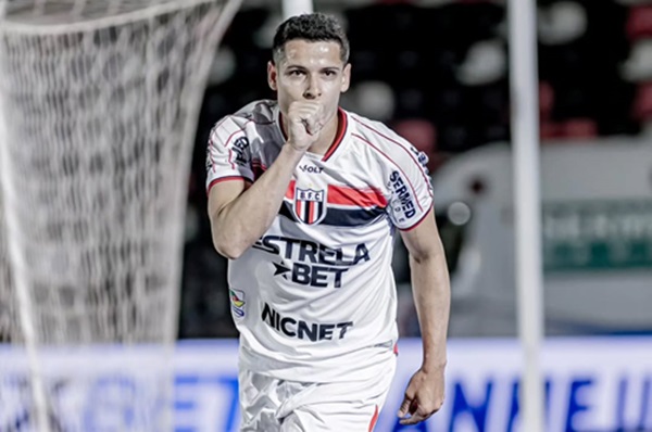 Fotografia de meio corpo do jogador de futebol Matheus Barbosa comemorando um gol pelo Botafogo-SP. Ele veste o uniforme branco com listras horizontais vermelhas e pretas no peito e o patrocínio "Estrela Bet". O jogador está com o punho fechado próximo à boca, em um gesto de silêncio ou beijo, olhando diretamente para a câmera. Ao fundo, vê-se a rede do gol e as arquibancadas desfocadas.