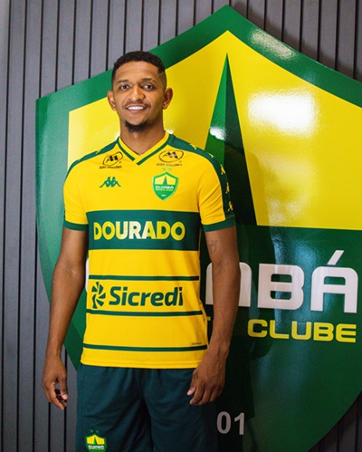Fotografia do atacante Hernandes sorridente, posando à frente de um grande escudo do Cuiabá Esporte Clube. Ele veste o uniforme oficial do time: uma camisa amarela vibrante com listras horizontais verdes e detalhes em verde na gola e mangas, exibindo os patrocinadores "Dourado" e "Sicredi". Ao fundo, o escudo gigante é verde e amarelo, posado contra uma parede de ripas verticais cinzas.