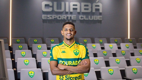 Fotografia de plano médio do atacante Hernandes, agora com os braços cruzados e um sorriso confiante. Ele está no centro de uma sala de imprensa ou auditório. Ao fundo, as poltronas cinzas possuem o escudo do Cuiabá bordado nos encostos. No topo da parede cinza ao fundo, destaca-se o letreiro metálico iluminado: "CUIABÁ ESPORTE CLUBE". A iluminação é suave, com faixas de luz verticais nas laterais.