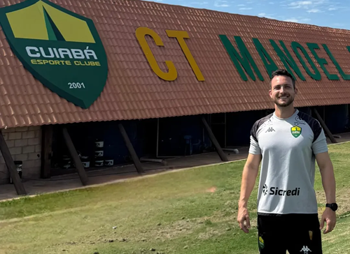Lucas Barezani posa sorrindo em frente ao prédio do CT Manoel Dresch, centro de treinamento do Cuiabá Esporte Clube, vestindo camisa oficial de treino do clube.
