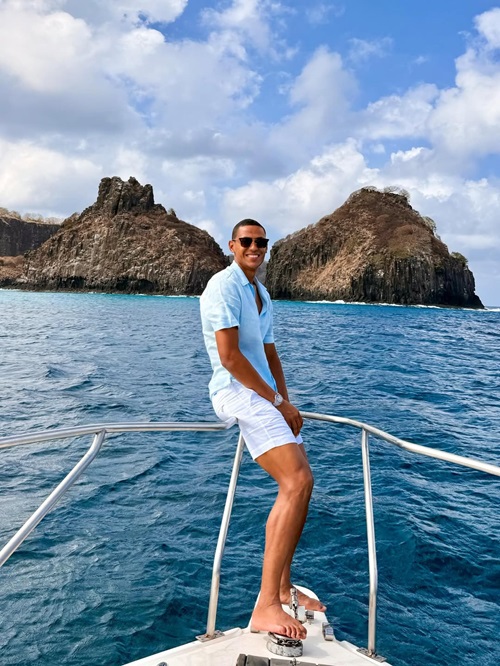 "Fotografia colorida do jogador Bruno Alves em um passeio de barco em Fernando de Noronha. Ele é um homem pardo, de sorriso aberto e óculos de sol escuros, vestindo uma camisa de linho azul clara de mangas curtas e shorts brancos. Ele está sentado na proa de um barco branco sobre o mar azul vibrante. Ao fundo, destacam-se as formações rochosas icônicas da ilha sob um céu com nuvens brancas."
