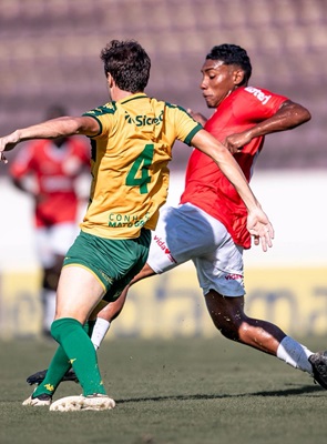 Lance de disputa de bola durante jogo da Copinha. Um jogador do Cuiabá, vestindo camisa amarela número 4 e calções verdes, está de costas em movimento defensivo contra um adversário que veste camisa vermelha e calção branco. A ação acontece em um gramado ensolarado com arquibancadas ao fundo.