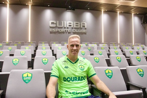 Fotografia do goleiro João Carlos sentado em uma das poltronas de uma sala de imprensa. Ele veste o uniforme verde-limão do Cuiabá e olha para a câmera com um sorriso amigável. As poltronas ao redor são cinzas com o brasão do clube. No topo da parede cinza ao fundo, destaca-se o nome do time em relevo sob iluminação focada.
