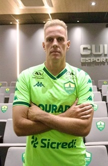 Fotografia em plano médio do goleiro João Carlos, do Cuiabá Esporte Clube. Ele é um homem branco, loiro, e está de pé com os braços cruzados e expressão séria. Veste o uniforme de goleiro na cor verde-limão com detalhes em verde escuro e patrocínios. Ao fundo, uma sala de imprensa com poltronas cinzas que levam o escudo do time e, na parede ao fundo, o letreiro "Cuiabá Esporte Clube".