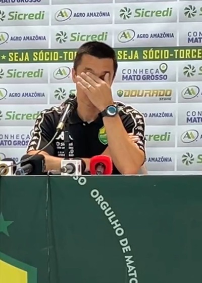 Fotografia de Eduardo Barros, integrante da comissão técnica do Cuiabá Esporte Clube, durante uma entrevista. Ele veste uma camisa polo preta do time e está à frente de um painel com logotipos de patrocinadores como Sicredi e Agro Amazônia.