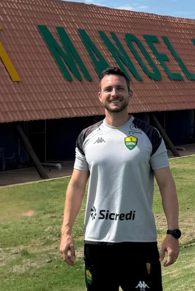 Lucas Barezani posa sorrindo em frente ao prédio do CT Manoel Dresch, centro de treinamento do Cuiabá Esporte Clube, vestindo camisa oficial de treino do clube.