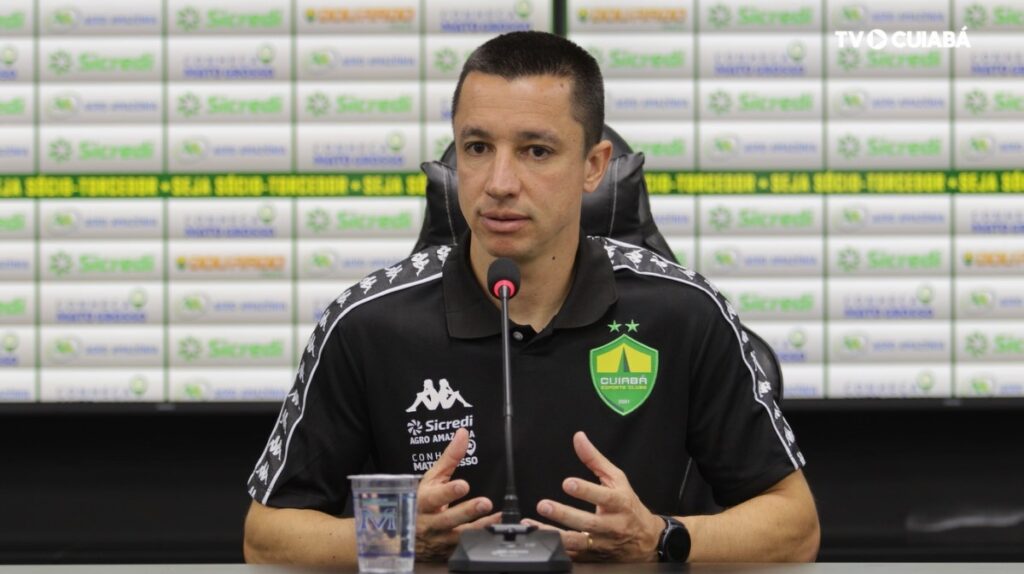 Fotografia de Eduardo Barros, integrante da comissão técnica do Cuiabá Esporte Clube, durante uma entrevista. Ele veste uma camisa polo preta do time e está à frente de um painel com logotipos de patrocinadores como Sicredi e Agro Amazônia.