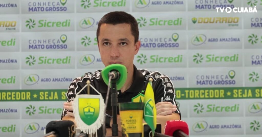 Fotografia de Eduardo Barros, integrante da comissão técnica do Cuiabá Esporte Clube, durante uma entrevista. Ele veste uma camisa polo preta do time e está à frente de um painel com logotipos de patrocinadores como Sicredi e Agro Amazônia.
